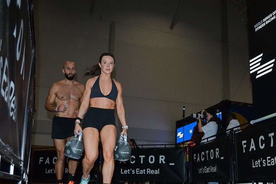 Alana carrying a kettlebell in each hand during her HYROX race, her teammate beside her
